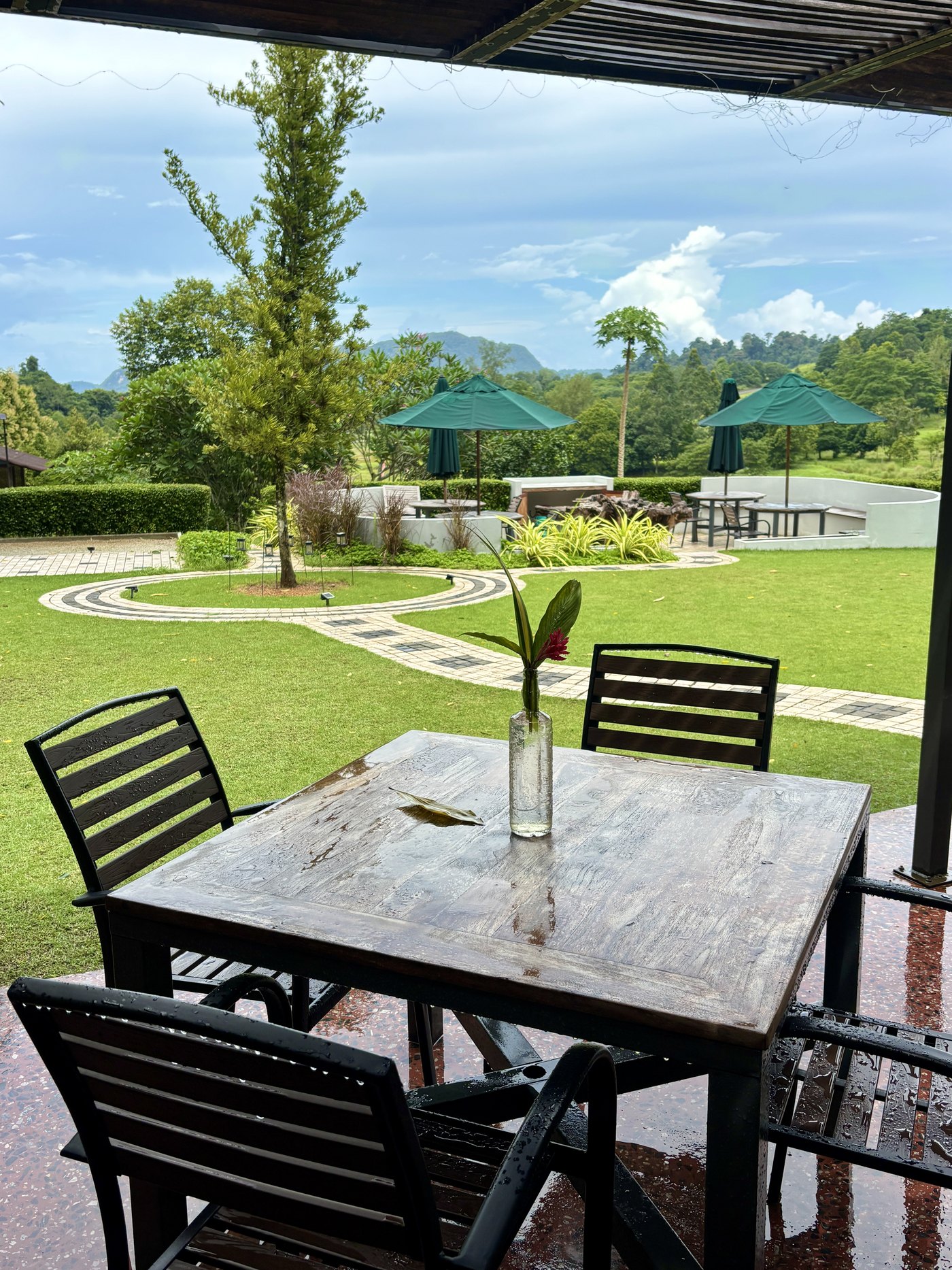 An empty timber table and slatted chairs under the roof, looking out across a mown lawn with garden umbrellas to trees and hills.
