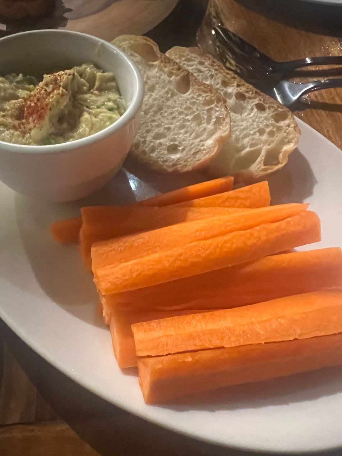 A bowl of hummus dusted with paprika, next to sliced baguette and carrot sticks on a white plate.