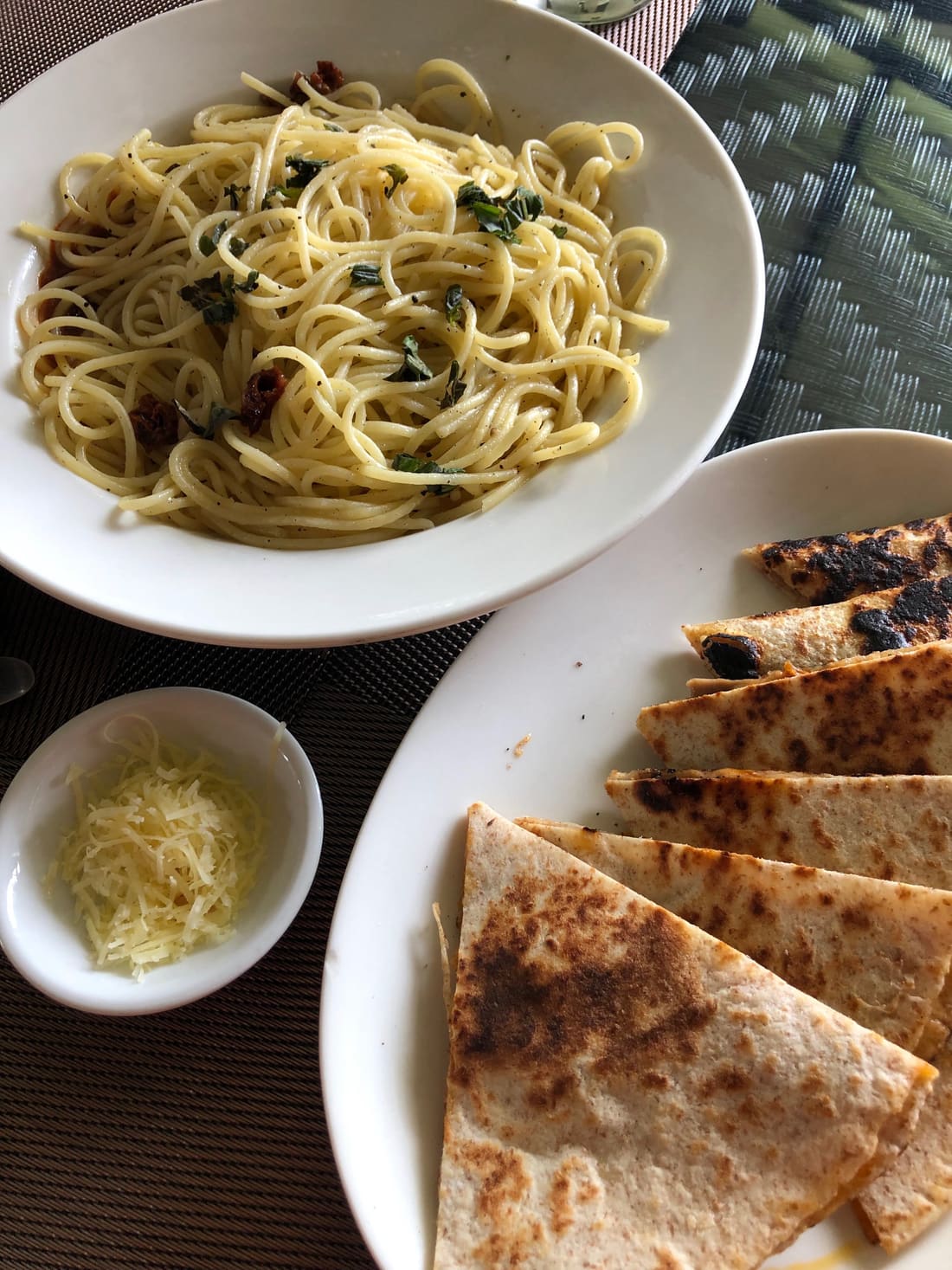 A plate of spaghetti aglio olio with herbs, with a small bowl of grated cheese beside it.