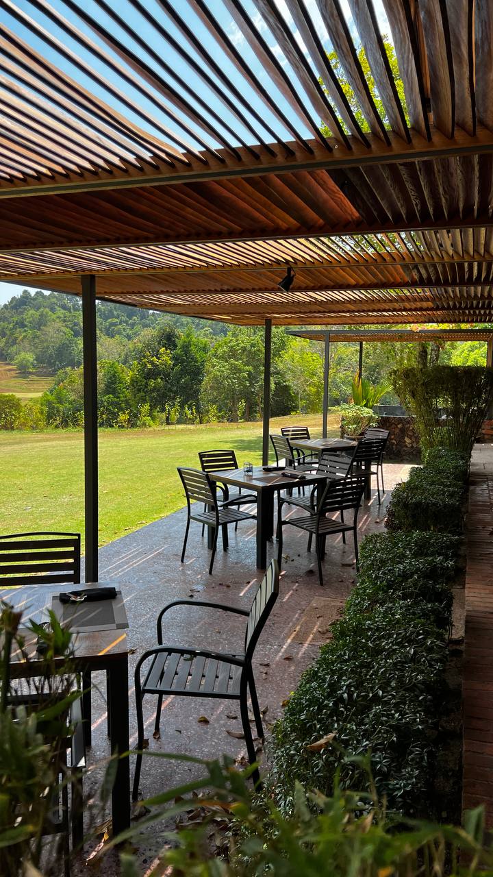 Outdoor tables under a slatted timber roof, looking across a mown lawn to trees and hills.